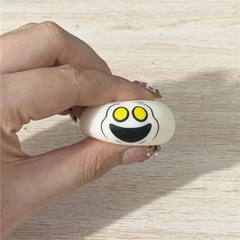A hand squishing a needoh ball mini squishkin with cartoon ghost face with yellow eyes and a black smile, against a light wood background.