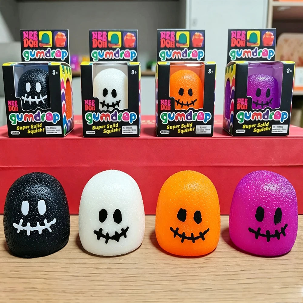 Four Halloween-themed squeeze squishies in ghost shapes displayed in front of their retail packaging. The squishies are black, white, orange, and purple, each with a spooky stitched face.